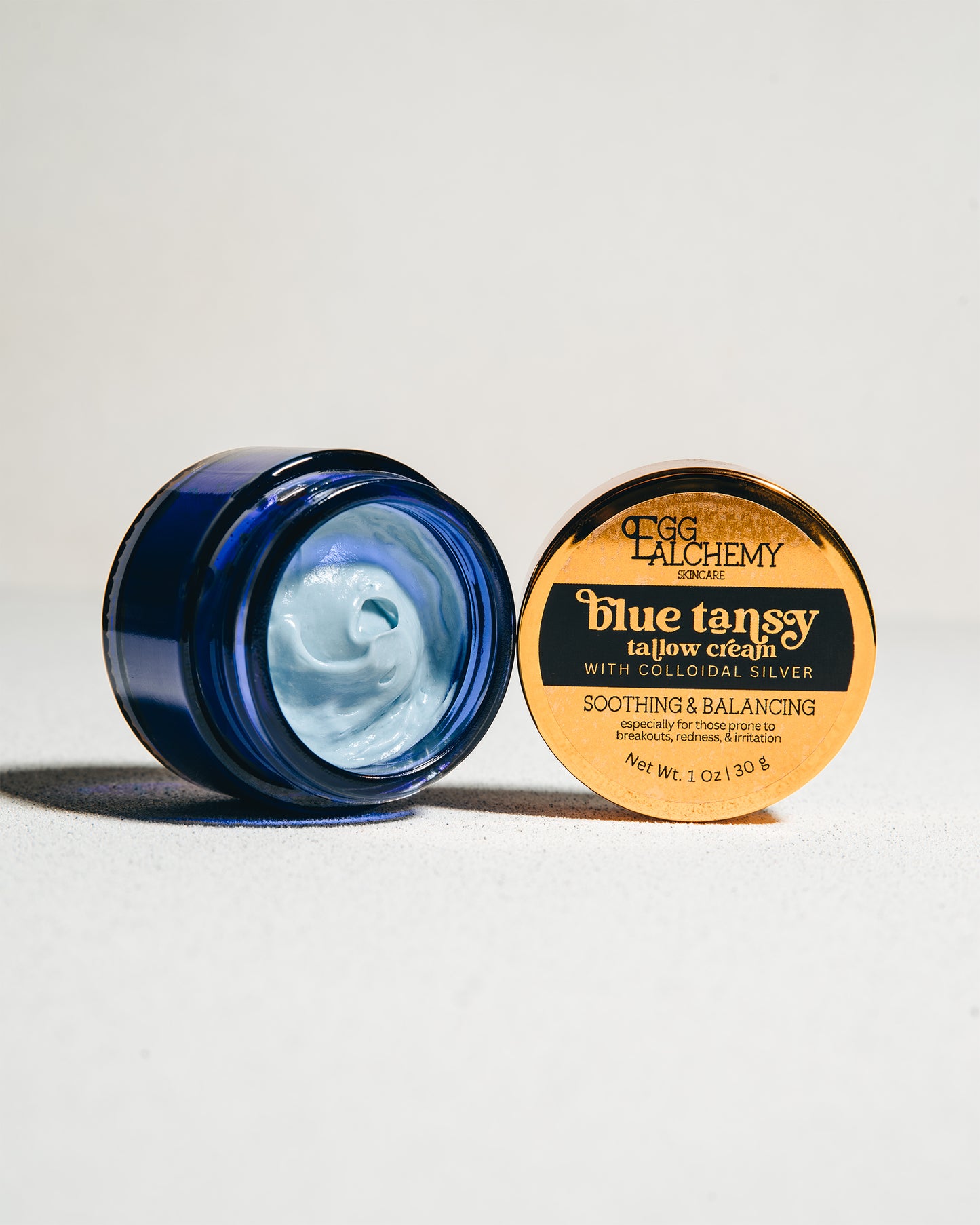 A blue glass jar sits open on its side, filled with Blue Tansy Tallow Cream with Colloidal Silver.  A shiny, gold lid stands on its side next to it.
